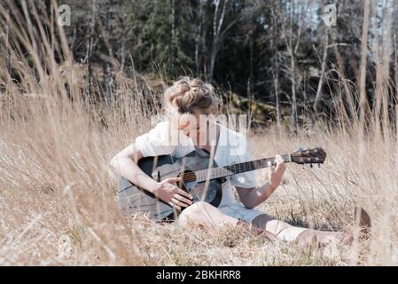 woman sat in a field happily playing guitar in summer Stock Photo