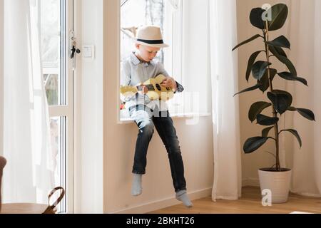 young boy sat on a window ledge at home playing ukulele Stock Photo