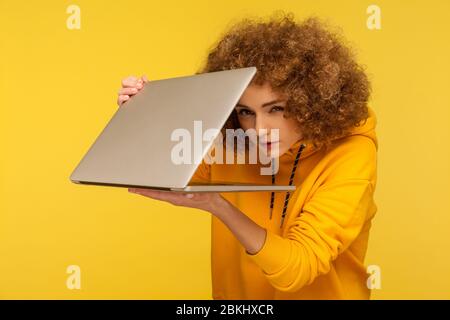 Woman with curly hair in urban style hoody peeking out half-closed laptop with sly suspicious look, spying and knowing secret information, using inter Stock Photo