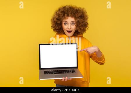 Shocked, surprised woman with fluffy curly hair in urban style hoody pointing at empty laptop screen and looking with amazement, showing mock up, blan Stock Photo