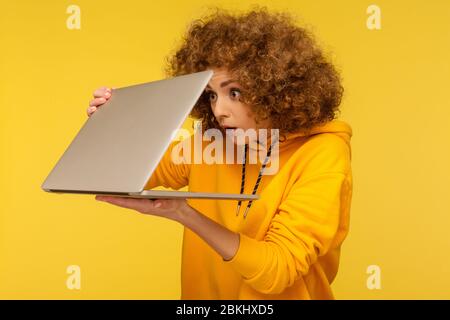 Shocked frightened woman with curly hair in urban style hoody looking furtively at half closed laptop screen, upset scared of error, software failure. Stock Photo