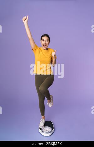 Full length of a cheerful young pretty sportswoman standing on scales isolated over violet background, celebrating Stock Photo