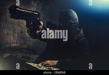 Cyber ​​criminal hacker in a dark room with low light at the computer, threatens with a gun Stock Photo
