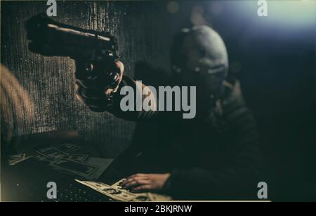 Cyber ​​criminal hacker in a dark room with low light at the computer, threatens with a gun Stock Photo