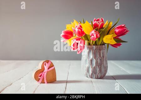 Bouquet of pink and yellow tulip flowers in a blue vase and heart shape gift box with satin ribbon bow on the white wooden table and grey background. Stock Photo