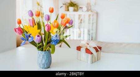 Fresh spring colorful bouquet of tulips, daffodils, irises in vase and gift box on white table with light classic design room background. Festive flow Stock Photo