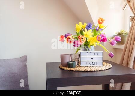 Everything for a calm stay at home. Lightbox with Stay home message, bouquet of fresh spring flowers and candles on the coffee table, and pillow sitti Stock Photo