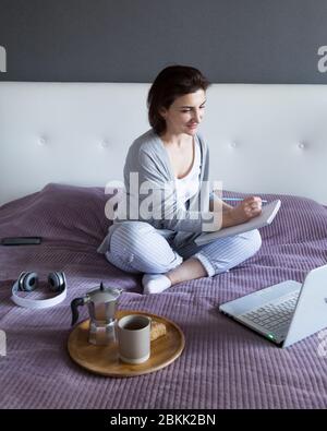 Young woman in grey working at home in her bedroom, freelance work with laptop and notepad. Distant education and training or home office concept Stock Photo