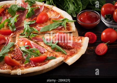 Pizza with Italian prosciutto, sun dried tomatoes and fresh arugula on dark background. Stock Photo