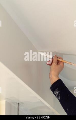 A portrait of a female hand and a dirty sleeve of a person holding a paint brush, painting in the corner made by the wall and the ceiling with white p Stock Photo