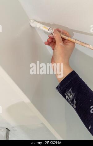 A portrait of a person painting the seam between the wall and ceiling of a room in a house with a paint brush in a white color. The person has a dirty Stock Photo