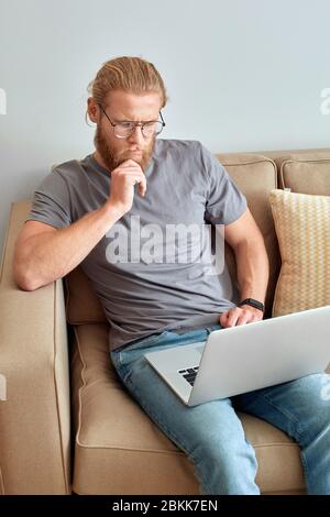Weekend at Home. Young man in glasses sitting on sofa working online on laptop touchign chin pensive Stock Photo