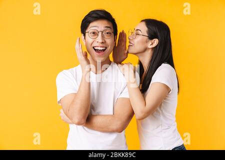 Image of smiling woman in eyeglasses whispering secret to surprised asian man isolated over yellow background Stock Photo