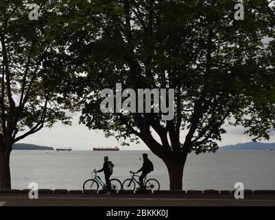 A pair of cyclists on ther bicycles are silhouetted as they take in views of English Bay and anchored freighters as they take advanatge of bike paths Stock Photo