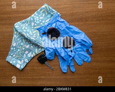 Gray cloth mask with flower and leaf pattern, car key, sunglasses, and blue disposable gloves on a wood table. Stock Photo