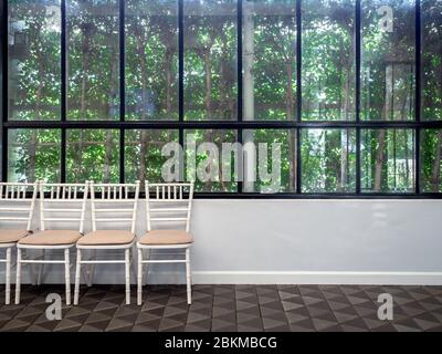 Row of clean white vintage wooden chairs with pads on modern stone tiles floor on white wall and glass window with green garden background. Stock Photo