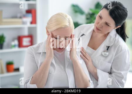 Blonde doctor holding her head and suffering from a headache, her colleague looking concerned Stock Photo