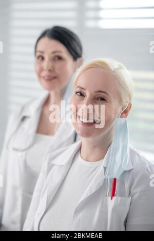 Two women in white coats with masks standing near window. Stock Photo