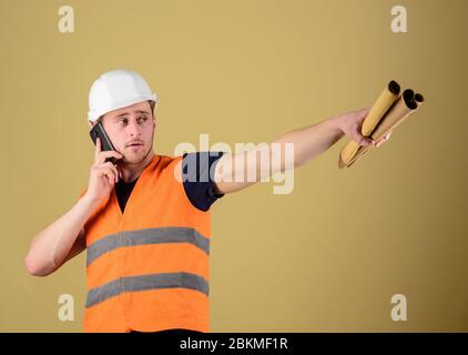 Man supervises construction on phone, ocher background. Engineer, architect on busy face speaks on phone, holds blueprints, pointing with finger, copy space. Supply of building materials concept Stock Photo