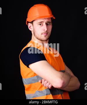 Strong builder concept. Builder in helmet posing confidently. Man in helmet, hard hat hold arms crossed on chest, black background. Worker, contractor, builder on serious face with muscular biceps Stock Photo
