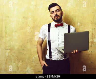 Manager with beard and confident face expression wearing white shirt, red bow and suspenders, holding laptop in hand, on beige background. Concept of work and business Stock Photo