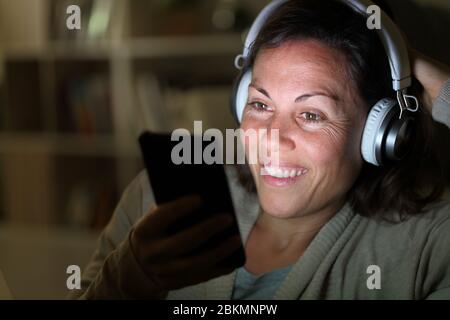 Happy adult woman listens music with headphones on smart phone with lighted screen sitting at night at home Stock Photo