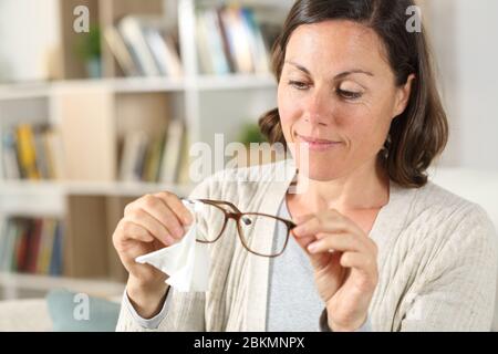 Happy adult woman cleaning eyeglasses with tissue paper sitting in the sofa at home Stock Photo