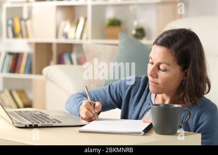 Adult woman with laptop writing notes on notebook sitting on the floor on a coffee table at home Stock Photo