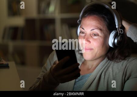 Happy adult woman with headphones listens to music on smart phone with lighted screen sitting at night at home Stock Photo