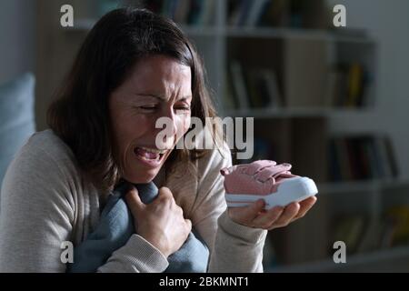 Sad mother crying missing her lost baby daughter holding shoe sitting at night at home Stock Photo