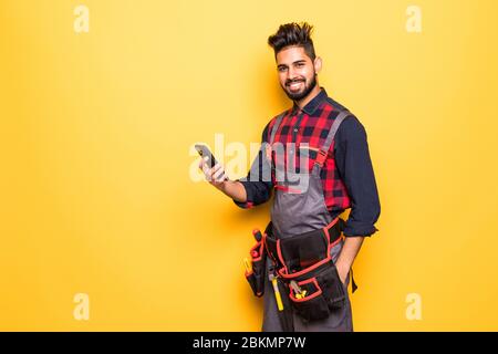 Portrait of young male construction worker with smart-phone wearing protective clothes, helmet and tool belt isolated Stock Photo