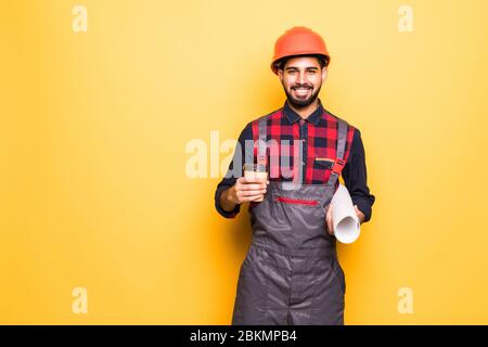 Smiling indian engineer in safety vest holding blueprints and coffee to go on yellow background Stock Photo