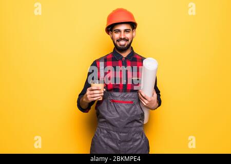 Smiling indian engineer in safety vest holding blueprints and coffee to go on yellow background Stock Photo