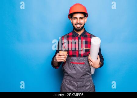 Smiling indian engineer in safety vest holding blueprints and coffee to go on blue background Stock Photo