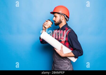 Smiling indian engineer in safety vest holding blueprints and coffee to go on blue background Stock Photo