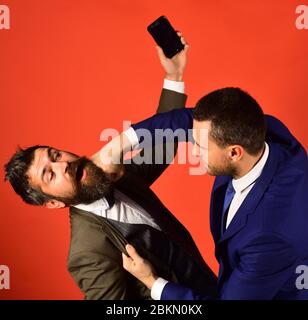Business talk and rivalry concept. Men with beards fight on red background. Businessman with surprised face holds mobile phone. Machos in classic suits talk on cell phone. Stock Photo