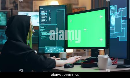 Female cyber terrorist wearing a hoodie working on computer with green screen. Stock Photo