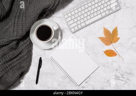 Autumn concept with cozy home office workspace. Marble desk with warm wool plaid, yellow leaves, coffee cup, keyboard, blank paper in notepad and pen Stock Photo