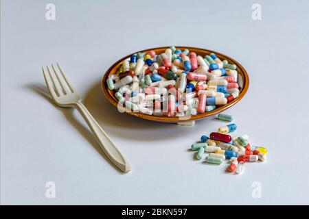 A small plate filled with differently colored medical capsules, some capsules separated, a white fork next to it, displayed on a white table Stock Photo