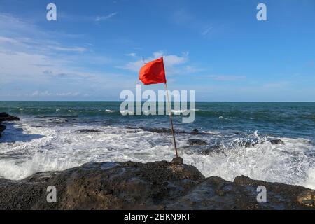 Red flag on a wooden flagpole flies on coast of Indian Ocean. Sign warning of danger of big breakers. Rock cliff at Sacred Balinese temple Tanah Lot. Stock Photo