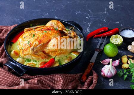 Thai coconut milk braised chicken and baby potatoes with chili, kaffir lime leaves in a black braiser on a kitchen table with meat fork and ingredient Stock Photo