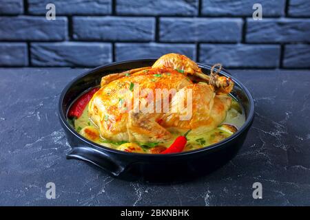 close-up of pot roasted coconut chicken with baby potato in a black ceramic braiser on a concrete table with a brick wall at the background, horizonta Stock Photo