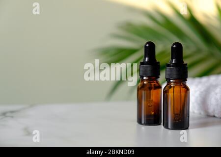 Two amber glass bottles for serum or essential oil on marble table, green palm leaf on the background. Brown dropper bottles with natural organic cosm