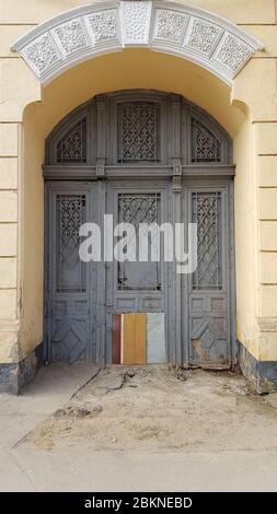 Damaged gates in arch of historic building with yellow wall and white ornate patterns. Doors protected by lattice with arabic ornament. Old entrance n