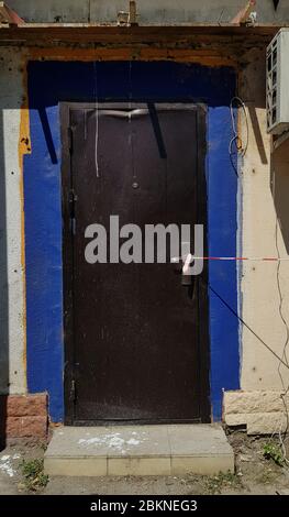 Dirty metal door of building under repair construction and closed by striped caution tape. Blue painted wall of house with forbidden entrance Stock Photo