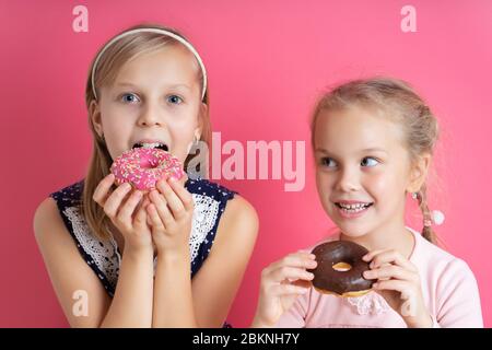 Two blonde sisters in colorful dresses. They are eating tasty donuts with sprinkle and chocolate-glazed. Posing on pink background Stock Photo