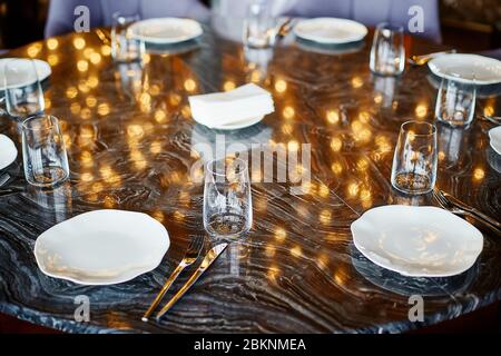 Eight platуs, forks, knives and glasses standing on a black marble table in which golden lights are reflected Stock Photo