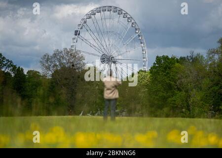 05 May 2020, Saxony-Anhalt, Magdeburg: A man is standing in the Rotehornpark on a flowering meadow in front of a big wheel. At present, the operators are working out a concept to comply with the distance rules. At the base, a snack bar has already opened and sold diner. However, eating is not permitted within a radius of 50 metres. The Ferris wheel had been set up in the past weeks and is operated by the Boos brothers from Magdeburg. Actually the ride should have been operated at other fairs at the moment. But they had been cancelled due to the spread of the corona virus. Now the Ferris wheel Stock Photo