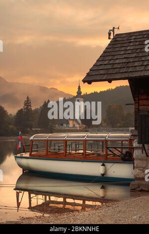 Electric motorboat for sightseeing tour of lake Bohinj in Slovenia, Church of St. John the Baptist and old stone bridge in background Stock Photo