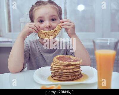 Funny little girl holds in front of the face a piece of pancake in the form of a smile. Stock Photo